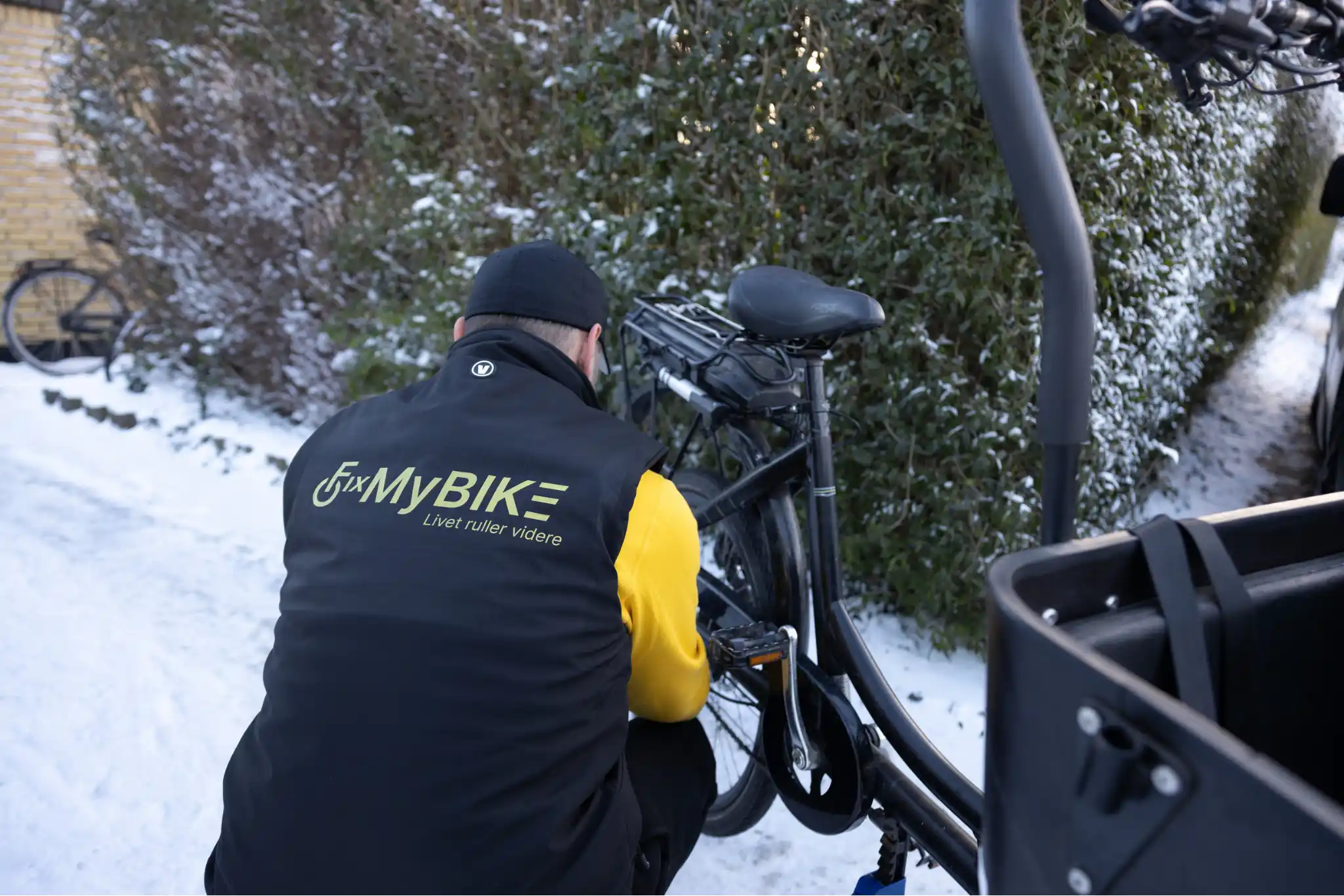 Cykelmekaniker i FixMyBike-uniform arbejder på en sort ladcykel i sneklædt indkørsel med hæk i baggrunden.