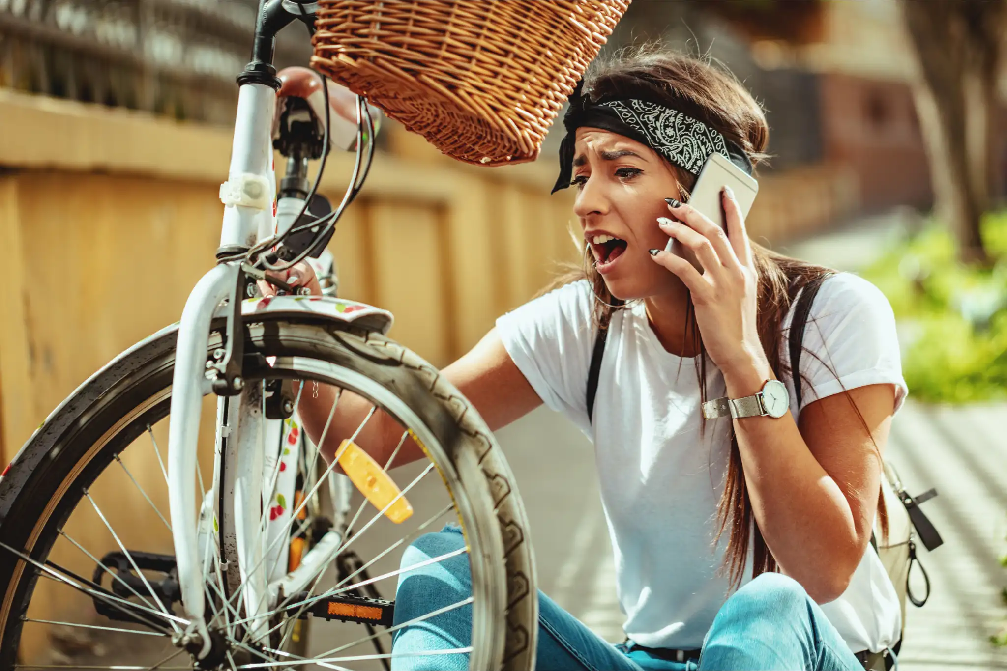 Frustreret kvinde med telefon i hånden sidder ved sin punkterede cykel og kigger på forhjulet.