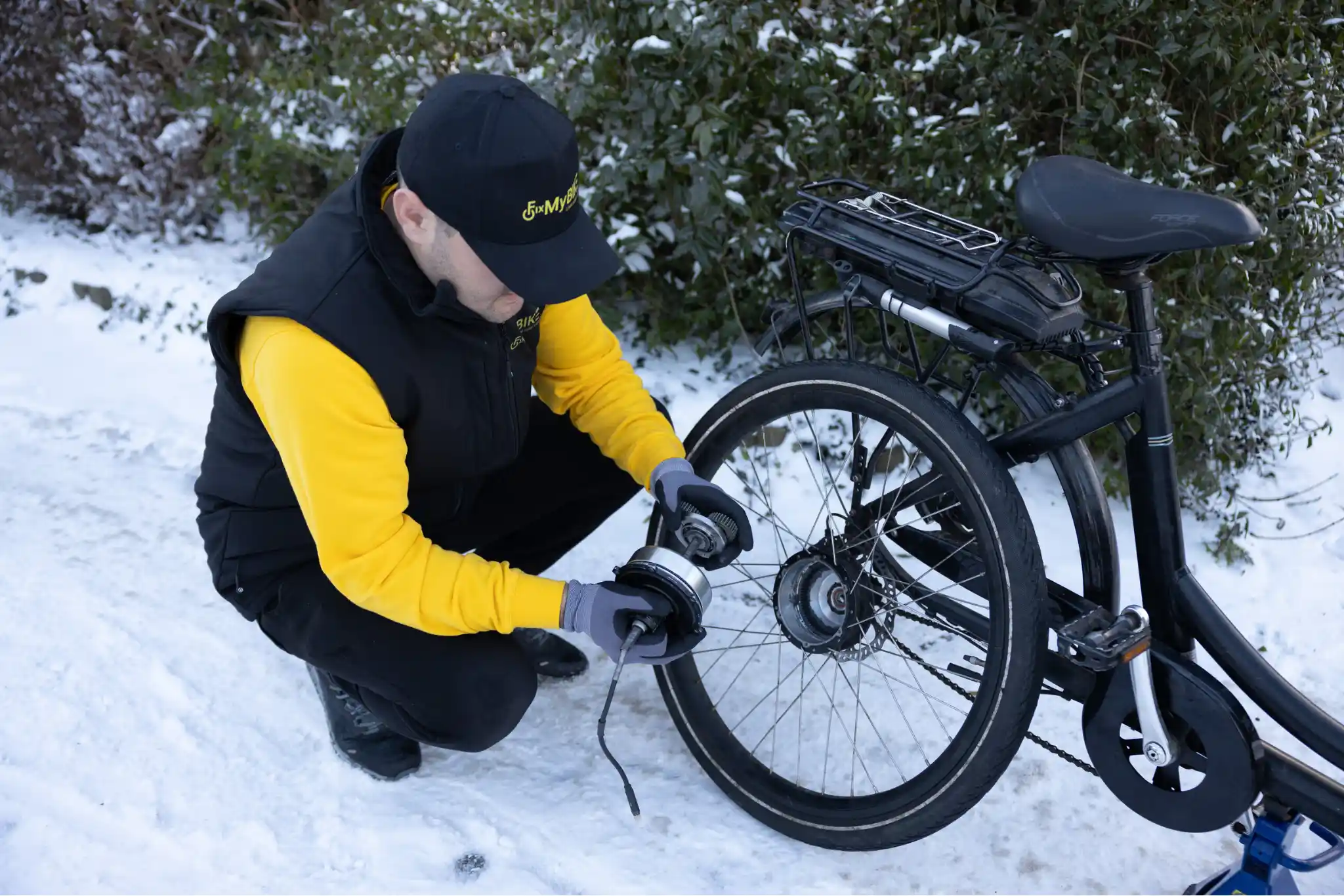Mekaniker fra FixMyBike arbejder på baghjulets elmotor på en sort ladcykel i sneklædt indkørsel.