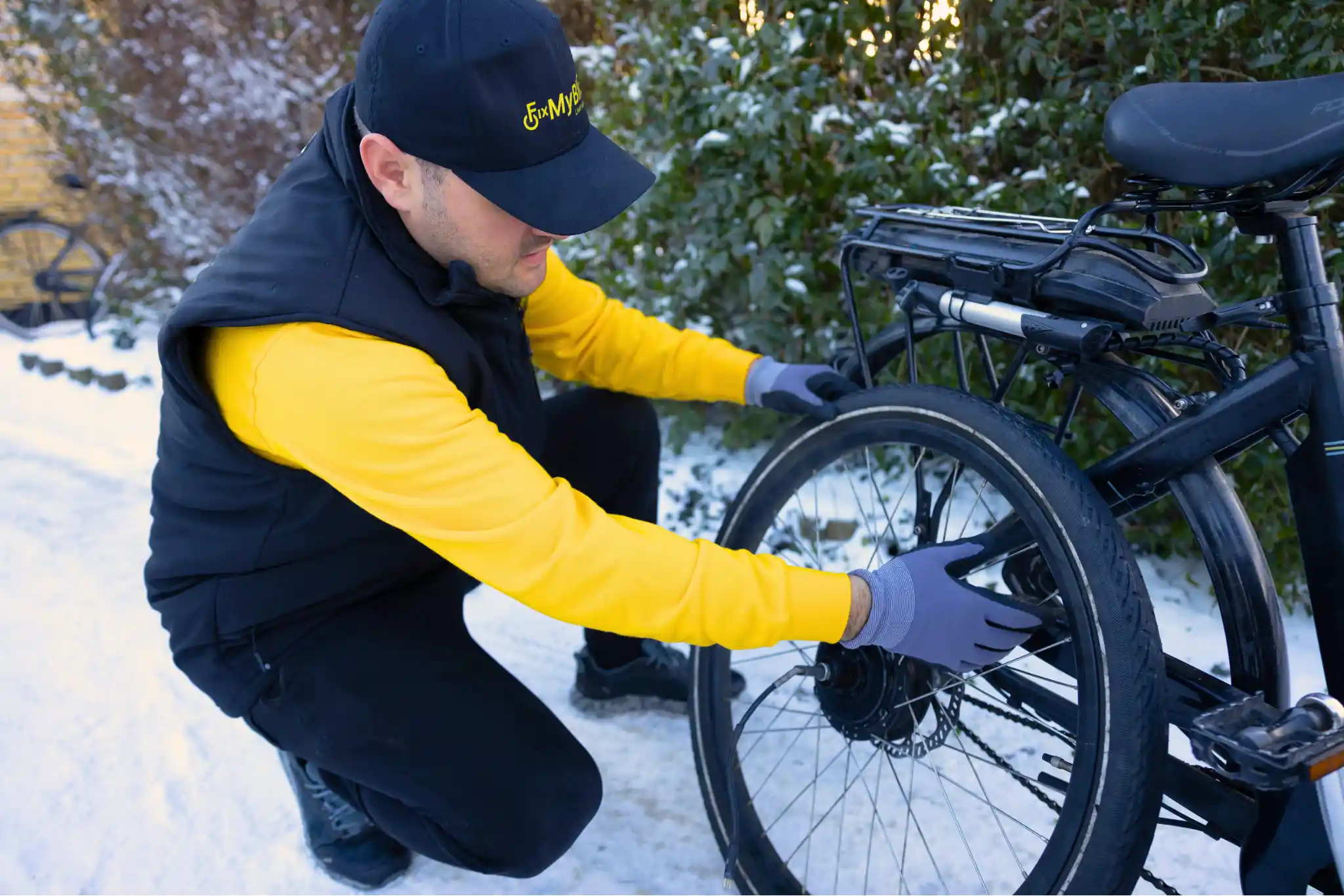 Cykelmekaniker fra FixMyBike monterer baghjul med motor på sort ladcykel i sneklædt indkørsel.