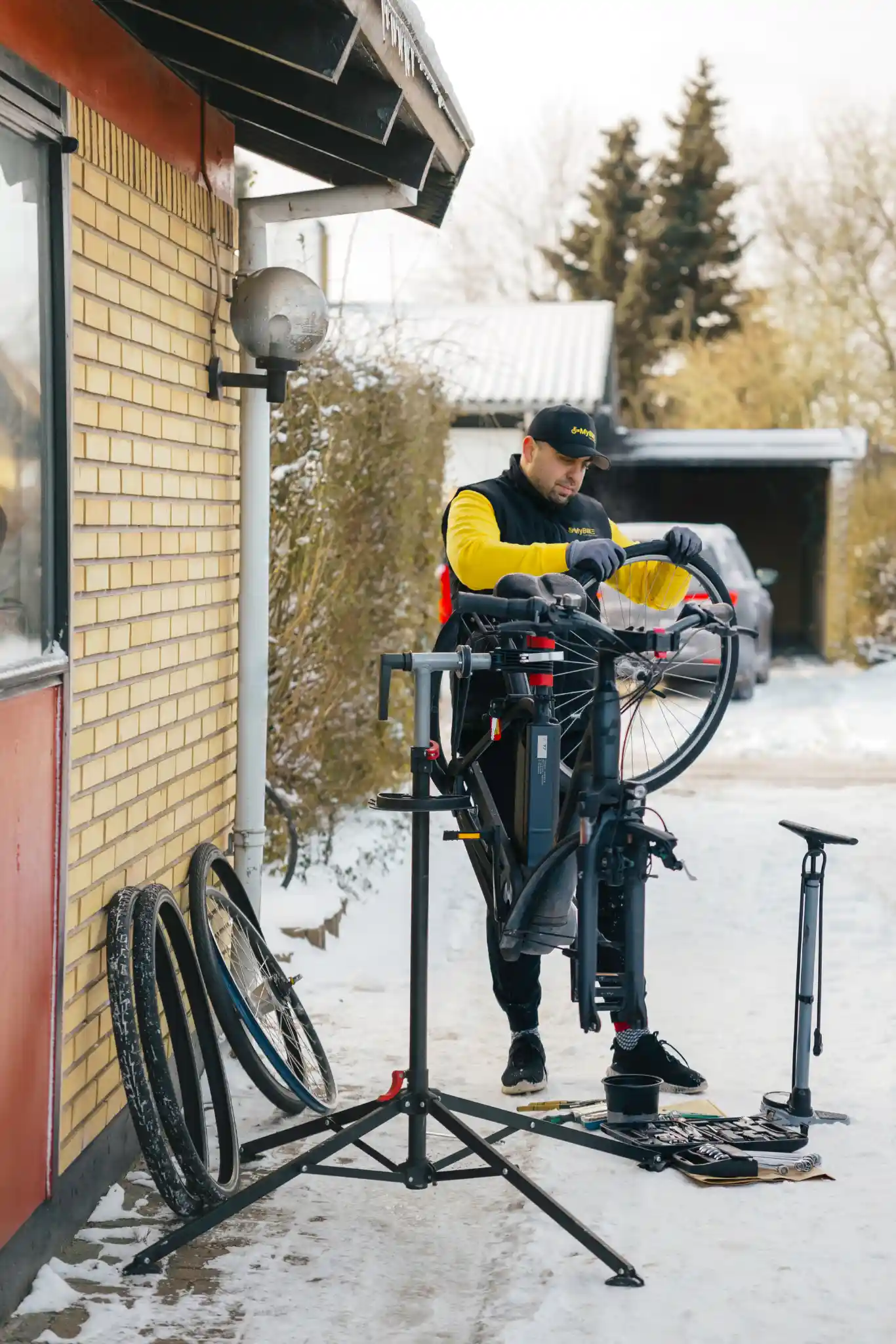 Cykelmekaniker fra FixMyBike arbejder udendørs i snevejr med at udskifte slange på elcykel, omgivet af værktøj og løse dæk.