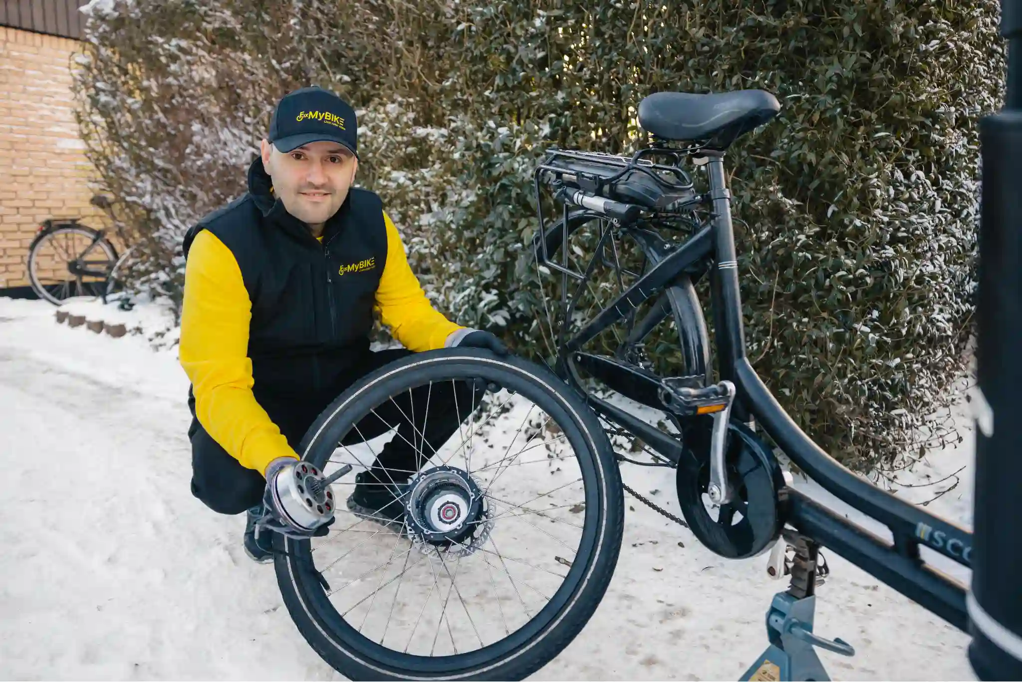 Cykelmekaniker fra FixMyBike holder baghjulet med elmotor til en sort elcykel under reparation i sneklædt indkørsel.