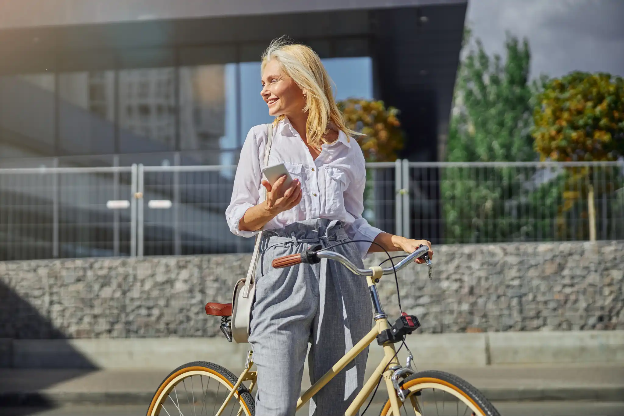 Smilende kvinde i lysegråt tøj står med mobil og beige cykel foran en moderne bygning i solskinsvejr.