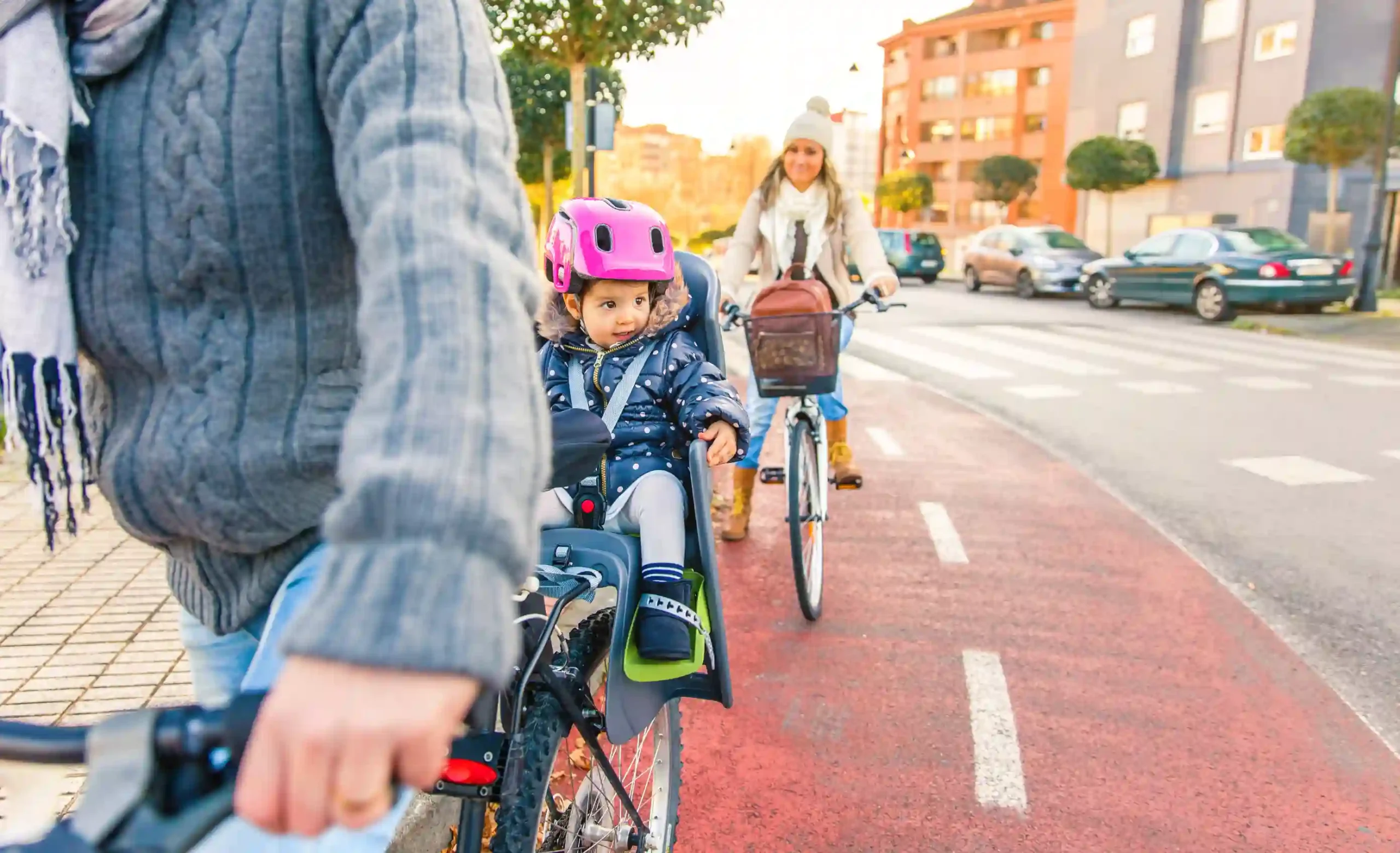Familie cykler på cykelsti; forælder i grå sweater cykler med lille barn i barnesæde og pink hjelm, mens en kvinde følger efter på hvid cykel.