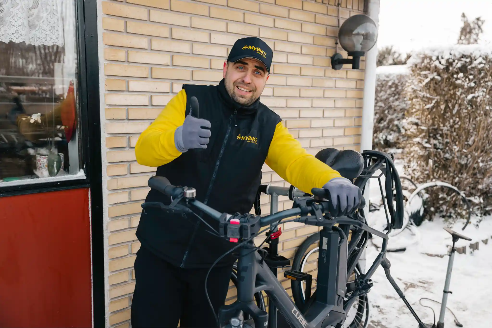 Smilende cykelmekaniker fra FixMyBike giver thumbs up ved siden af en elcykel efter reparation i sneklædt indkørsel.