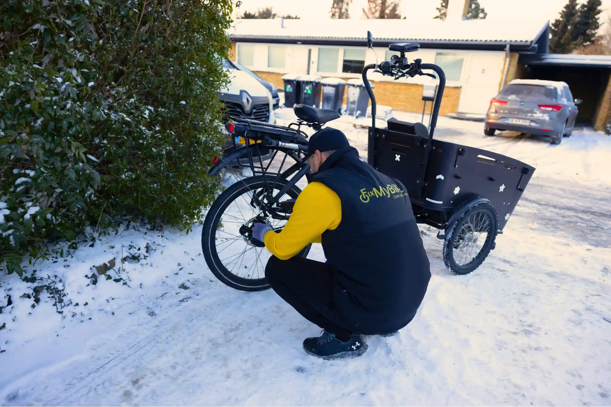 Cykelmekaniker fra FixMyBike reparerer baghjul på sort ladcykel i en sneklædt indkørsel foran en villa.