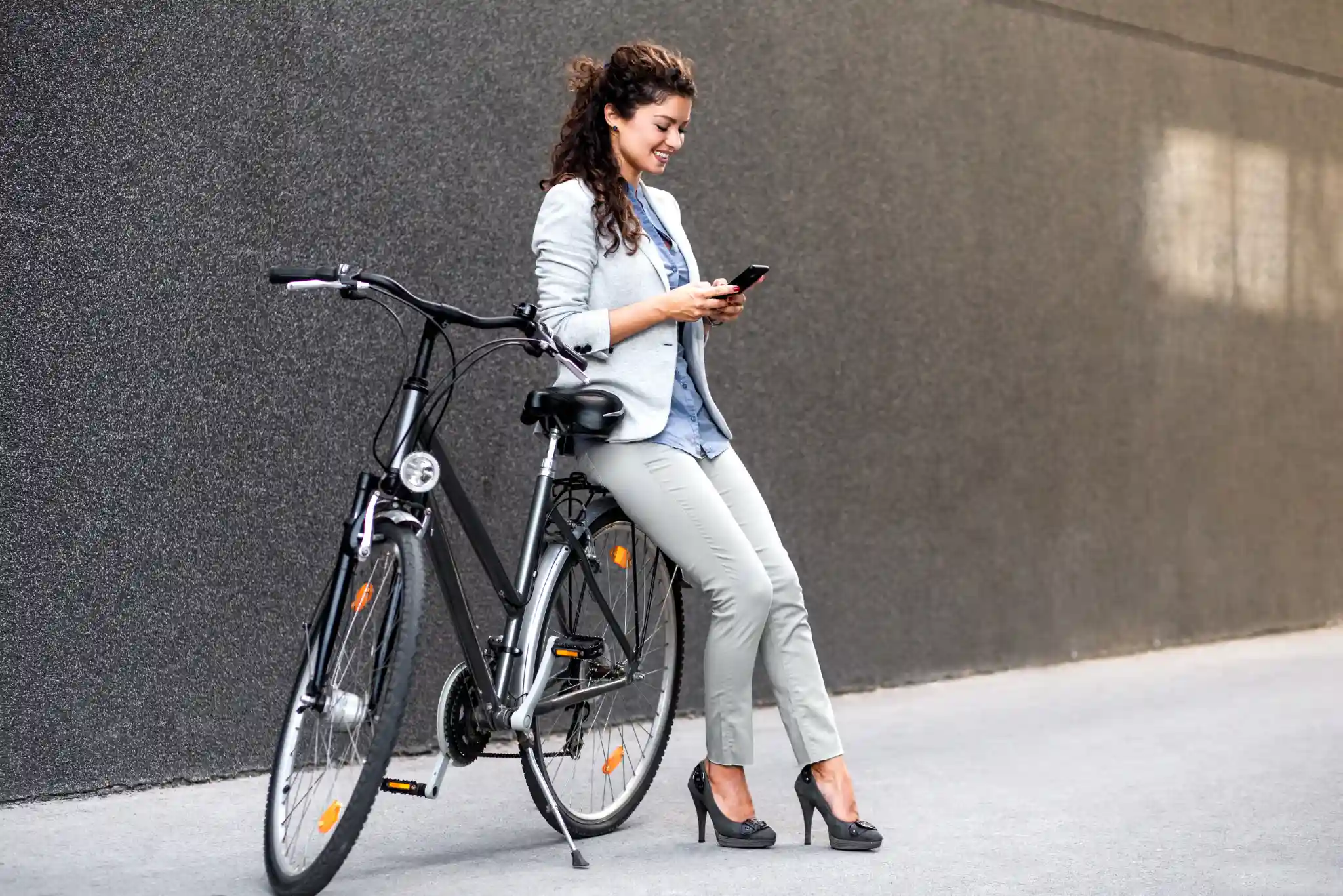 Smilende kvinde i business-tøj læner sig op ad en sort cykel, mens hun bestiller cykelservice på sin mobiltelefon.