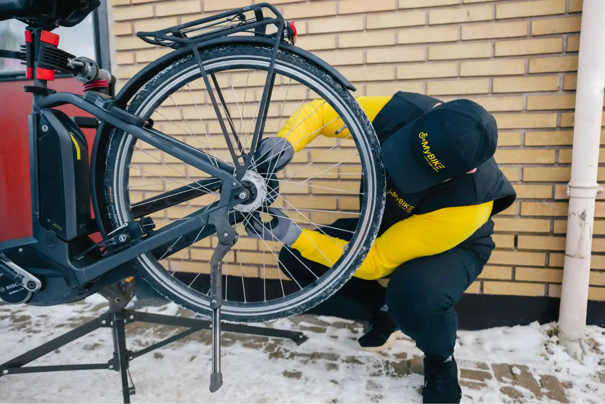 Cykelmekaniker fra FixMyBike efterspænder baghjul på elcykel med Bosch-motor foran murstensvæg i snevejr.