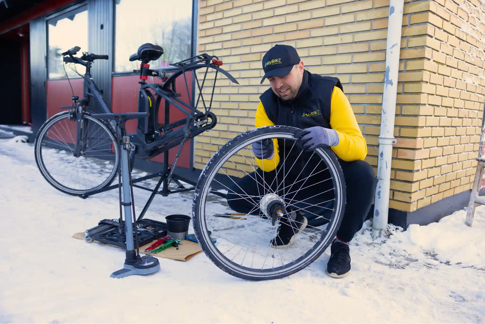 Smilende cykelmekaniker fra FixMyBike skifter slange på cykelhjul foran elcykel og værktøj i sneklædt indkørsel.